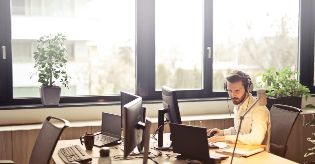 A businessman sits at a desk using multiple computers and a headset in a well-lit modern office.